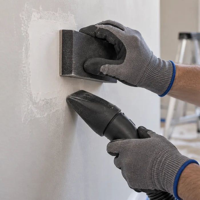 Illustration of gloved hands sanding a pale wall patch with a rectangular block and a vacuum nozzle below.