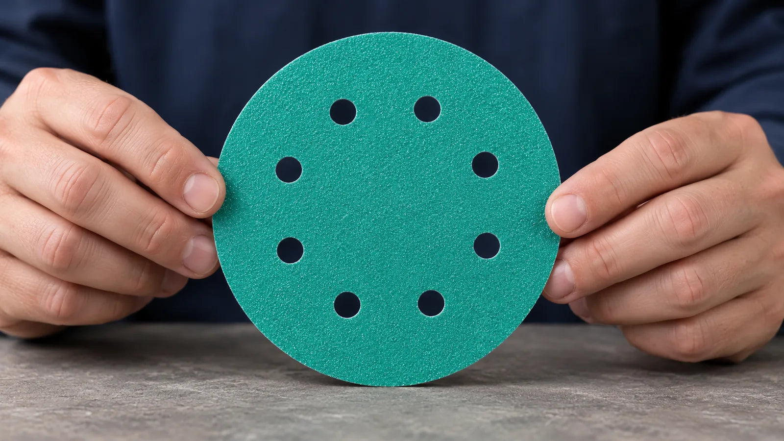 Two hands hold a green abrasive disc with eight visible holes for inspection.